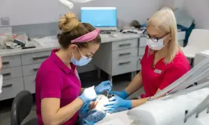 Dwie dentystki w maseczkach i rękawiczkach pracują przy pacjencie. Jedna z nich wykonuje zabieg, druga asystuje. Znieczulenie u dentysty działa szybko, by zabieg był komfortowy.