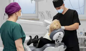 Dentysta zakłada szwy po wyrwaniu zęba. Pacjentka w fotelu, asystentka stoi obok.