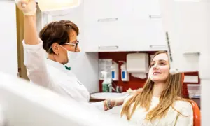 Dentystka w masce i rękawiczkach przygotowuje się do zabiegu, podczas gdy młoda kobieta w ciąży uśmiecha się, oczekując na wyrywanie zębów.