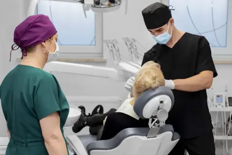 Dentysta zakłada szwy po wyrwaniu zęba. Pacjentka w fotelu, asystentka stoi obok.
