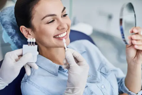 Uśmiechnięta kobieta porównuje odcień zębów z wzornikiem podczas zabiegu wybielania zębów u dentysty.