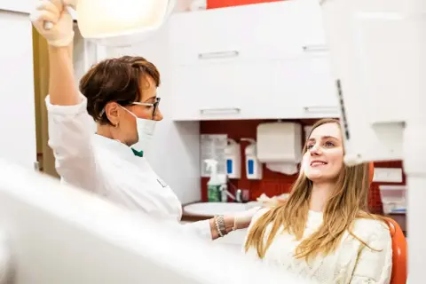 Dentystka w masce i rękawiczkach przygotowuje się do zabiegu, podczas gdy młoda kobieta w ciąży uśmiecha się, oczekując na wyrywanie zębów.