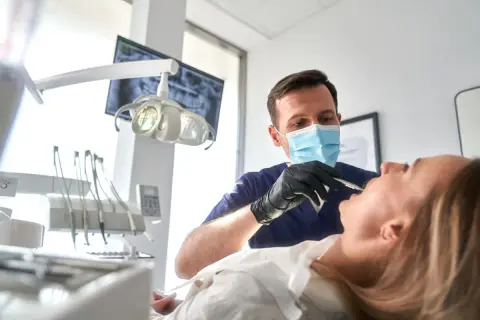 Dentysta w masce wykonuje zabieg szynowania zębów u pacjentki.