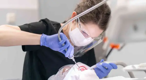 Dentystka w masce i rękawiczkach wykonuje zabieg. Leczenie zębów pod narkozą ryzyko jest minimalizowane przez profesjonalizm.