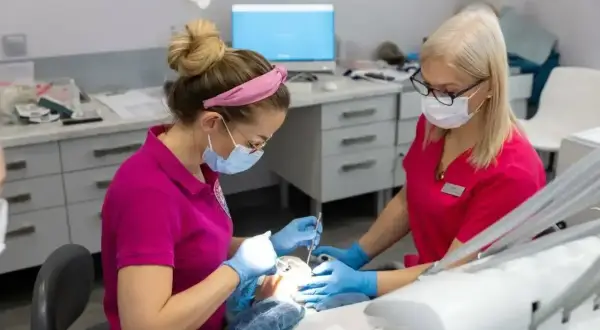 Dwie dentystki w maseczkach i rękawiczkach pracują przy pacjencie. Jedna z nich wykonuje zabieg, druga asystuje. Znieczulenie u dentysty działa szybko, by zabieg był komfortowy.