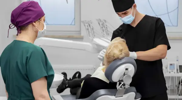 Dentysta zakłada szwy po wyrwaniu zęba. Pacjentka w fotelu, asystentka stoi obok.