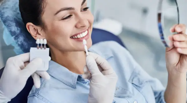 Uśmiechnięta kobieta porównuje odcień zębów z wzornikiem podczas zabiegu wybielania zębów u dentysty.