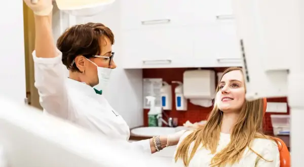 Dentystka w masce i rękawiczkach przygotowuje się do zabiegu, podczas gdy młoda kobieta w ciąży uśmiecha się, oczekując na wyrywanie zębów.
