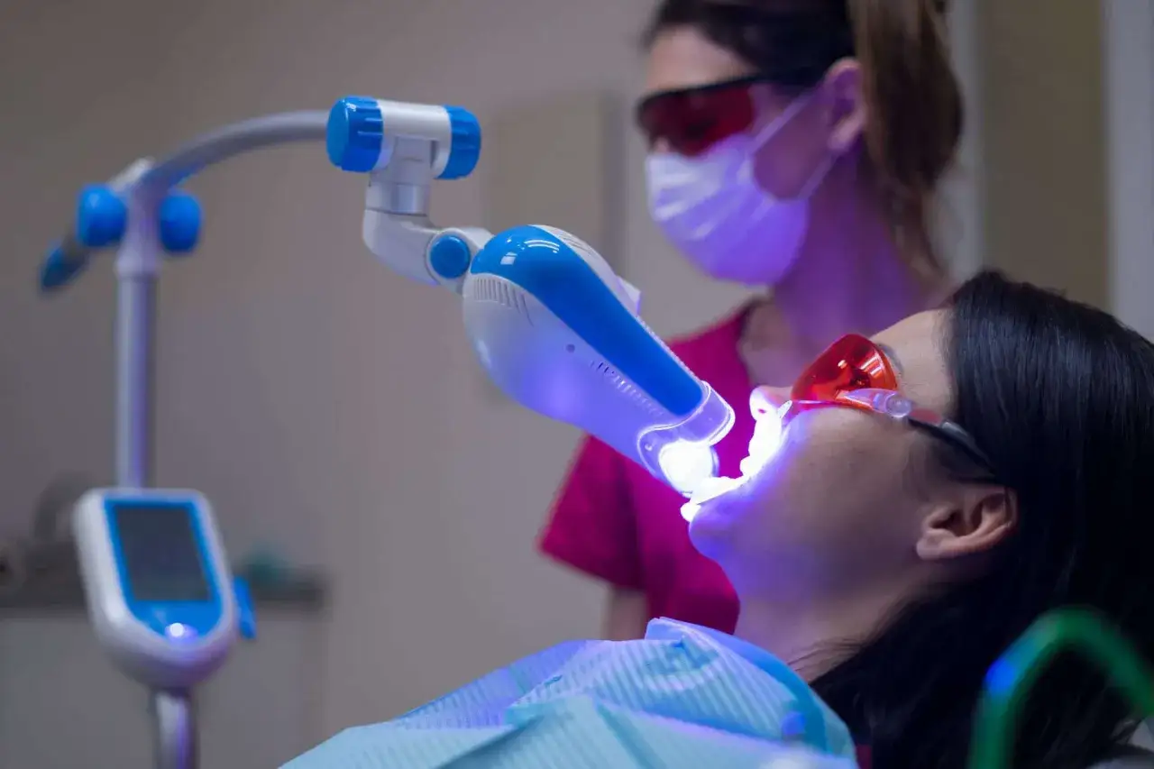 Pacjentka przechodzi zabieg laserowe wybielanie zębów. Dentystka w okularach ochronnych i masce nadzoruje proces.
