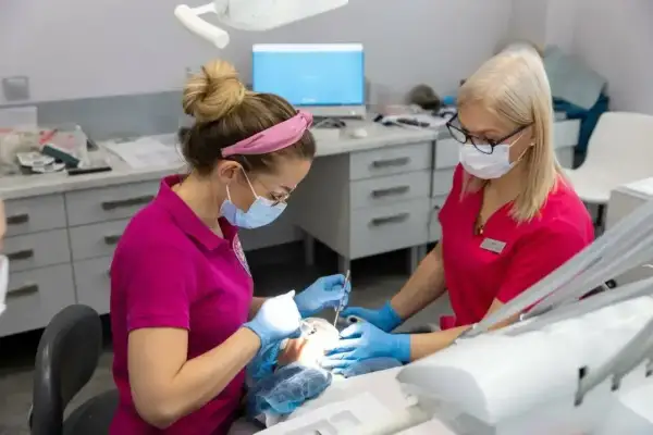 Dwie dentystki w maseczkach i rękawiczkach pracują przy pacjencie. Jedna z nich wykonuje zabieg, druga asystuje. Znieczulenie u dentysty działa szybko, by zabieg był komfortowy.