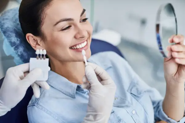 Uśmiechnięta kobieta porównuje odcień zębów z wzornikiem podczas zabiegu wybielania zębów u dentysty.