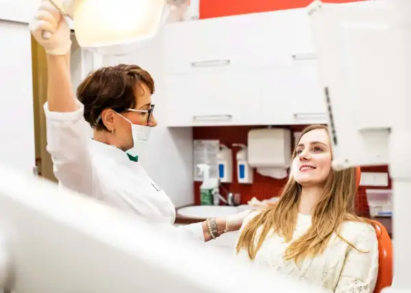 Dentystka w masce i rękawiczkach przygotowuje się do zabiegu, podczas gdy młoda kobieta w ciąży uśmiecha się, oczekując na wyrywanie zębów.