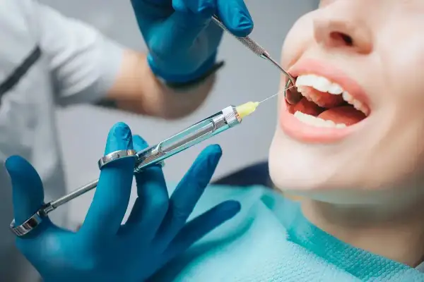 Dentysta podaje znieczulenie powierzchniowe. Pacjentka ma otwarte usta, a stomatolog używa strzykawki i narzędzia.